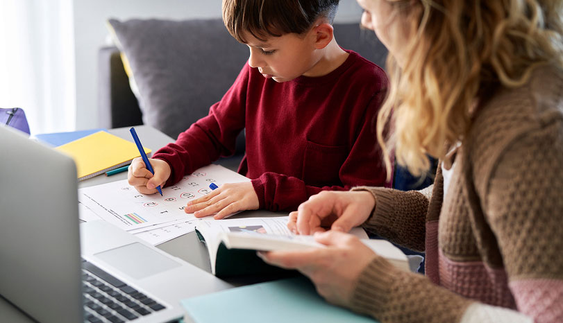 A young boy in a red sweater focuses on homework, writing with a blue pen. An adult beside him reads a book. A laptop and notebooks are on the table, conveying a learning atmosphere.