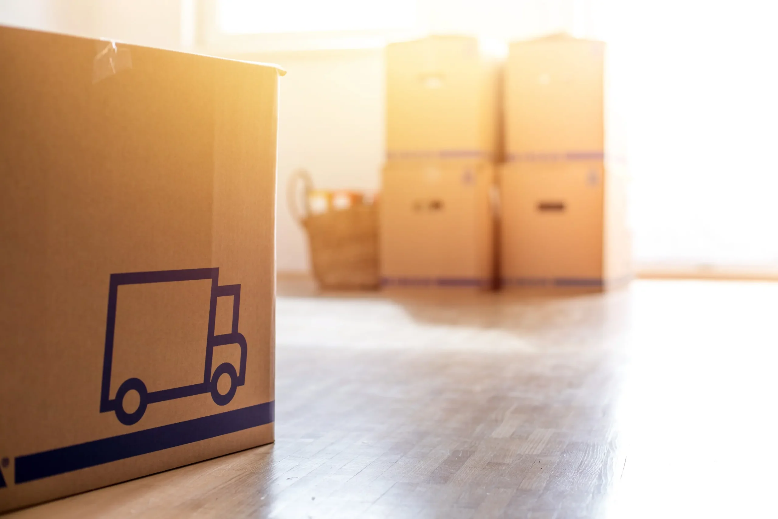 Cardboard boxes in a sunlit room suggest moving or packing. A large box with a truck icon is in focus, with several stacked boxes blurred in the background.