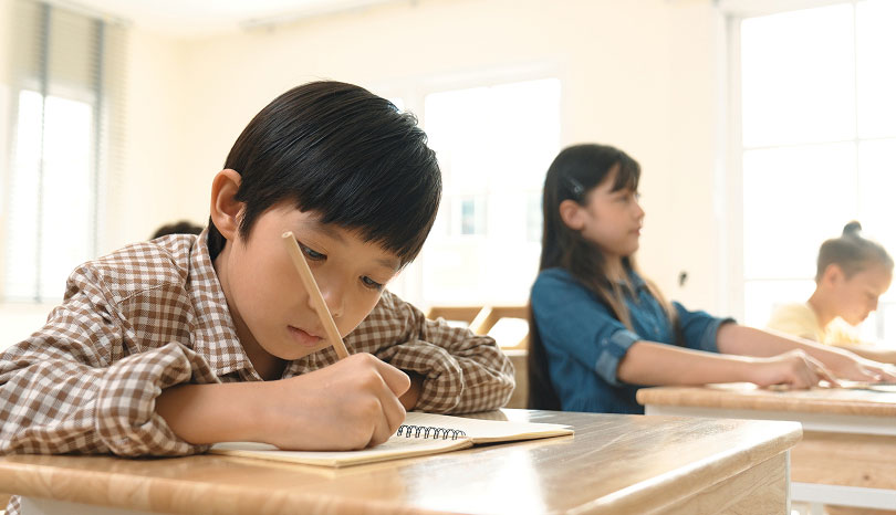 Young boy in a plaid shirt concentrates on writing in a notebook at a school desk. Behind him, two students, a girl and a boy, work quietly. Bright classroom.