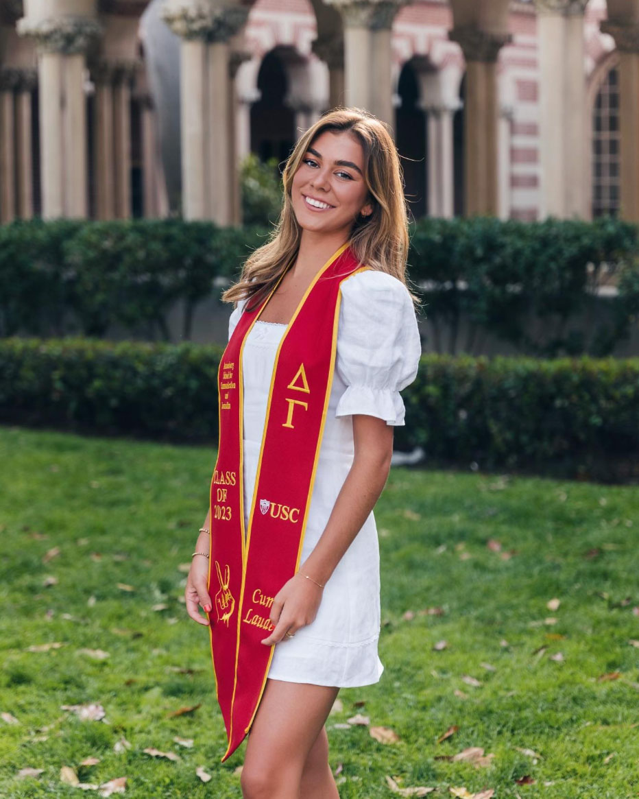 A young woman in a white dress stands smiling on a grassy area. She wears a red and gold graduation stole with Greek letters. The background shows an ornate building.