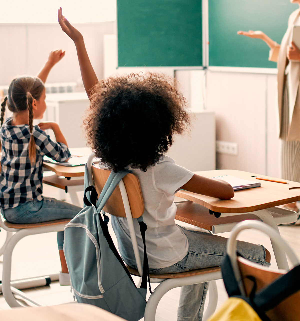 Two students sit at desks in a classroom, both raising their hands to respond. The setting feels engaging and educational, with a teacher gesturing at a chalkboard.
