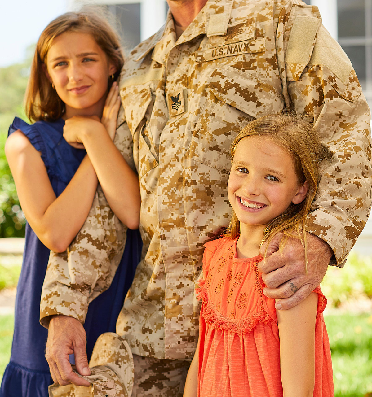 A Navy officer, wearing a camo uniform, lovingly embraces two smiling girls, one in blue and one in orange, against a sunny outdoor backdrop.