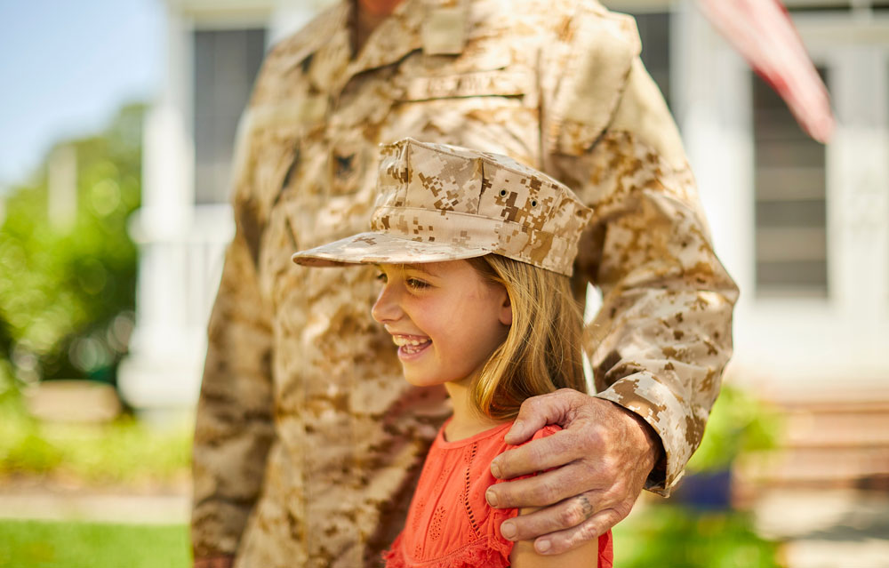 A smiling girl in a coral dress wears a military cap, hugged by a man in camo. The scene conveys warmth and pride in a sunlit outdoor setting.