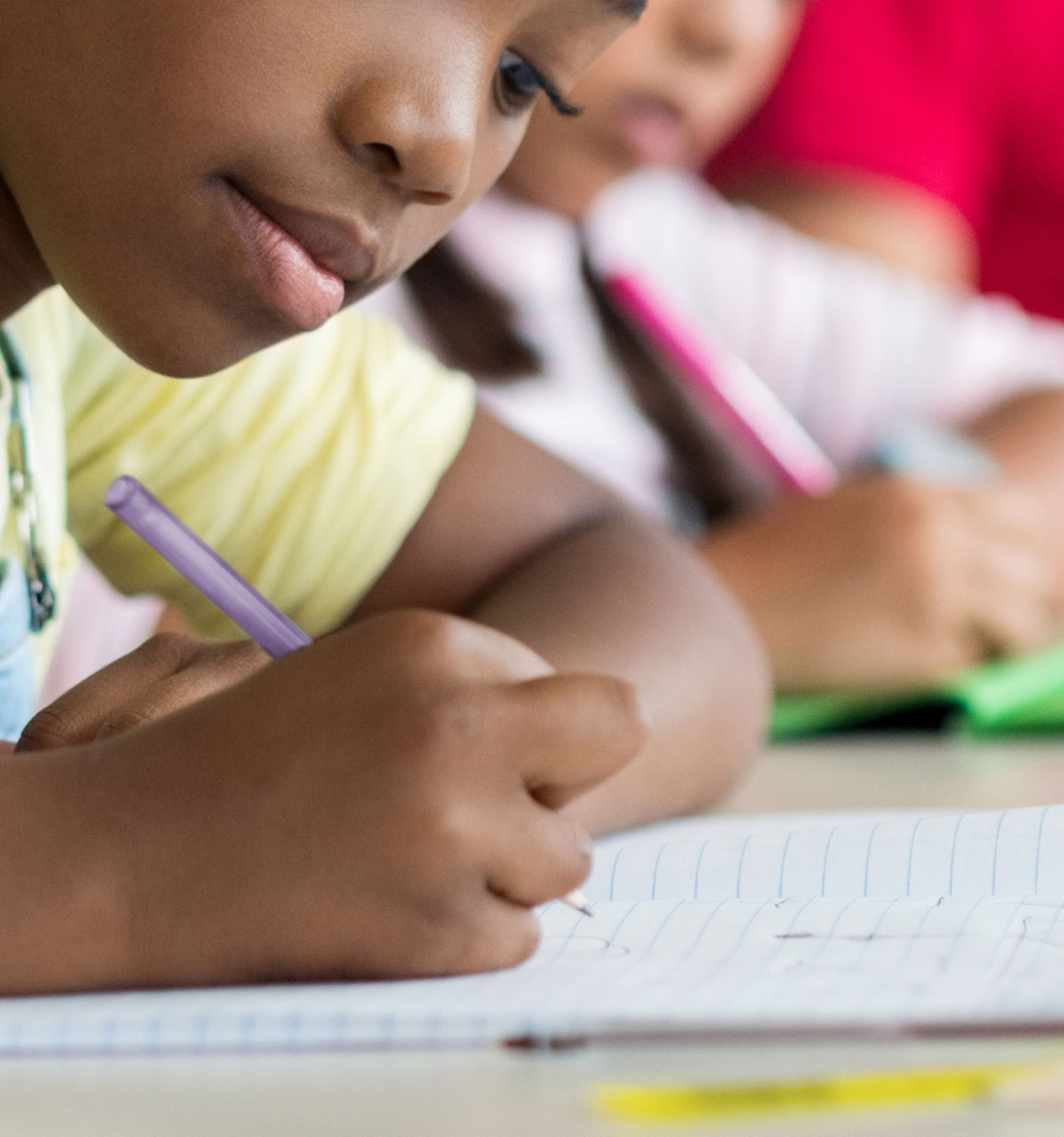 Young student writing intently with a purple pen on lined paper in a classroom. Others appear in the background, focused. The atmosphere is studious.