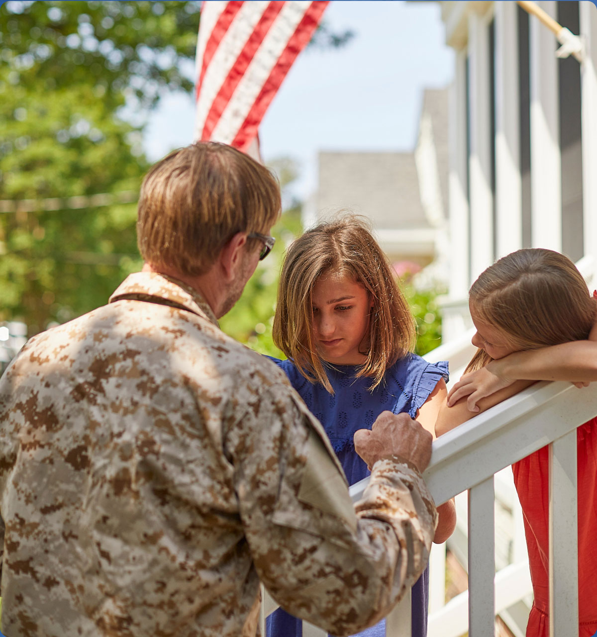 A man in a camo uniform interacts solemnly with two girls on a porch, one looking down, the other resting, with an American flag in the background.