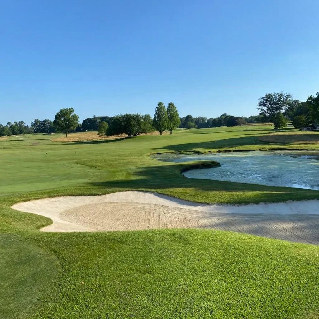 img-delaware-valor-classic A serene golf course under a clear blue sky displays lush green fairways, a sandy bunker, and a small pond, with trees lining the horizon.
