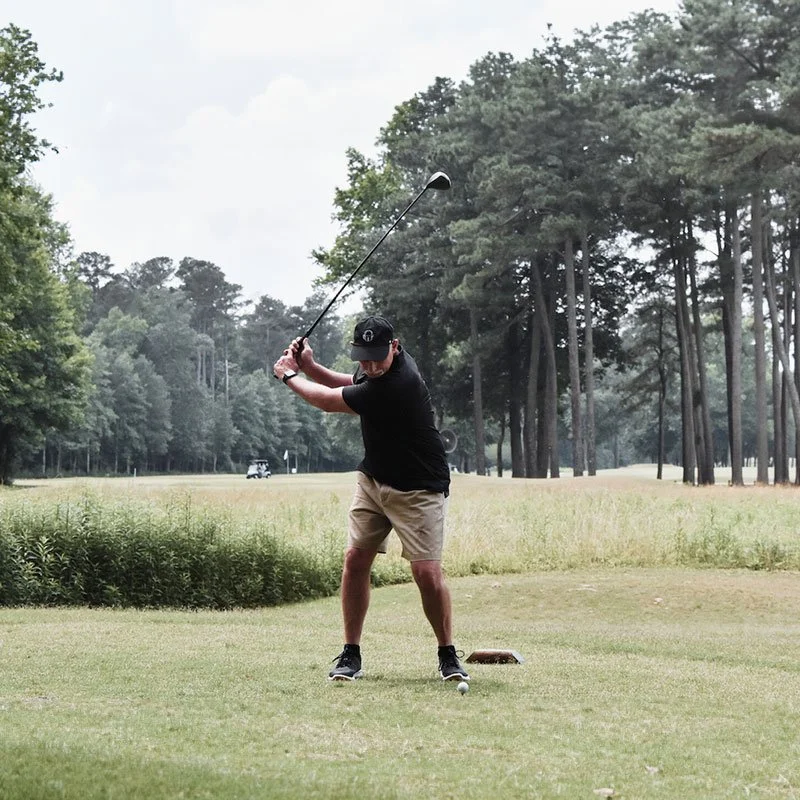 img-golf-valorclassic A golfer in shorts and a cap is captured mid-swing on a lush green course, surrounded by tall trees under an overcast sky. Calm and focused ambiance.